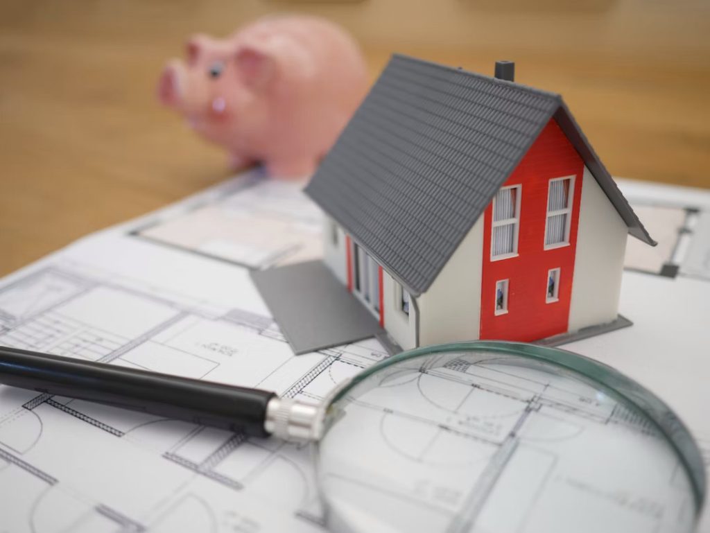magnifying glass, house model and piggy bank on top of blueprints